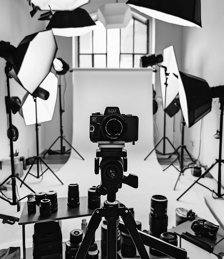 A black and white high-angle shot of a professional photography studio featuring a camera on a tripod, several softbox lights on stands, and multiple camera lenses on a side table.